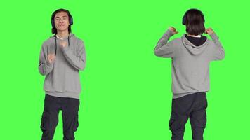 Positive guy does funky dance moves on camera, feeling groovy while he is listening to modern songs. Asian person smiling and enjoying music on headphones, dancing on greenscreen. video