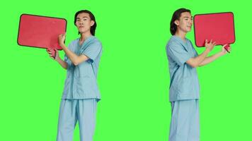 Medical nurse presents speech bubble in studio with greenscreen backdrop, holding cardboard icon with isolated copyspace. Healthcare worker showing small billboard sign on camera. video