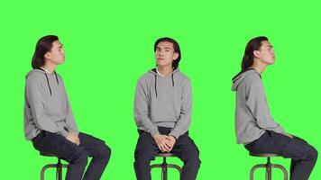 Young man sitting on chair and waiting for something in studio, looking at watch and searching for a thing against greenscreen backdrop. Male model with natural impatient expressions. video