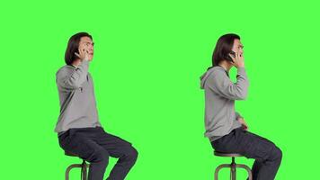 Modern guy making phone calls on camera, sitting on chair against greenscreen backdrop and having remote conversation. Young person being relaxed talking to friends, telephone line. video