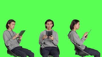 Asian man navigating internet on tablet in greenscreen studio, using mobile device to search through webpage online. Person sitting on chair and working on gadget, relaxed guy. video