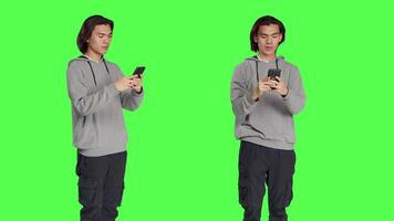 Guy texting messages on mobile phone standing against greenscreen backdrop, using social media app to chat remotely with friends. Asian joyful adult messaging on smartphone. video