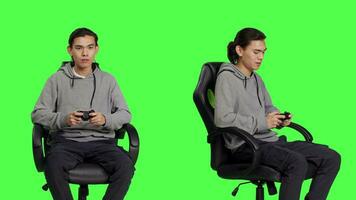 Asian guy using controller for gameplay, sitting on chair against greenscreen backdrop in studio. Young man enjoying online gaming plan with friends, gamer playing rpg video games.