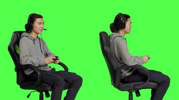 Cheerful person enjoys online gameplay, sitting on gaming chair over greenscreen isolated background in studio. Young asian guy having fun with competition, man playing video games.