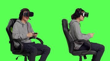 Gamer uses virtual reality for gameplay with mobile phone app, sitting on gaming chair in isolated greenscreen studio. Asian person playing multiplayer action video games with vr.