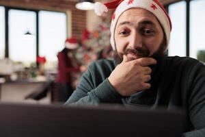 Smiling worker in santa hat rubbing chin while reading research report on laptop in office. Caucasian man company employee working on computer at christmas eve during winter holiday photo
