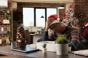 Depressed african american man in santa hat sitting at workplace desk after getting fired on christmas eve. Upset company employee dismissed from work during winter celebration photo