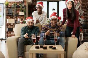 Happy coworkers group celebrating christmas together by playing video game in festive decorated office. Colleagues with gamepads having fun at new year corporate party in workplace photo