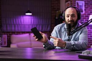 Portrait of tech vlogger reviewing newly released smartphone in rgb lights living room studio. Entertaining content creator filming technology channel episode using professional video tools photo