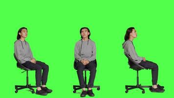 Young guy looks at wristwatch and waiting in studio, acting impatient while sitting on chair over full body greenscreen. Agitated person checking time on watch, trying to stay positive. video