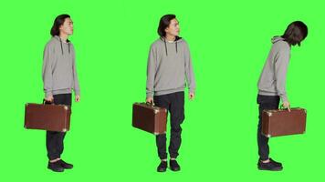 Young adult carries retro suitcase over full body greenscreen backdrop on camera, waiting impatiently for a thing. Asian model holding big suitcase while he checks time on wrist watch. video