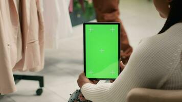 Käufer im zweite Hand Kleidung Geschäft halten Grün Bildschirm Tablette, versuchen zu entscheiden Was Kleider zu kaufen. Klient suchen beim Neu Sammlung von Kleider im Rabatt Geschäft mit Chroma Schlüssel Gerät video