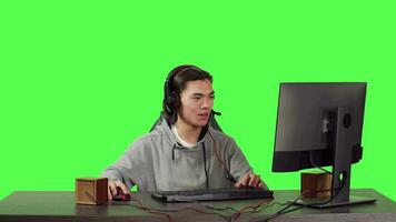 Masculin joueur à poste de travail pièces sur ordinateur personnel, ayant amusement avec en ligne rpg tournoi pour en ligne vidéo Jeux avec autre personnes. asiatique homme jeu à ordinateur plus de plein corps écran vert toile de fond. video