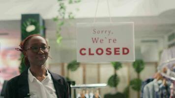 Portrait of cheerful BIPOC woman opening stylish fashion boutique, starting her shift in the morning. Employee turning closed sign on fast fashion clothing store into open one video