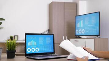 Man working at the desk in living room checking some data on computer screen. There is a big screen TV displaying the same data. Dolly slider 4K footage video