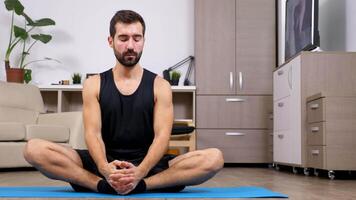 joven ajuste hombre haciendo yoga, el loto pose, en el piso de su departamento. él sitts en un azul estera con cerrado ojos. muñeca control deslizante 4k imágenes video