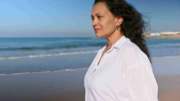 retrato de pelinegro medio Envejecido natural mujer en casual ropa, sonriente disfrutando su caminar solo en hermosa arenoso playa en soleado día, dejando huellas en un mojado arena lavado por atlántico olas video