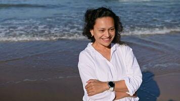 Close-up portrait of a beautiful middle aged woman in white shirt, standing with arms folded on the seashore, smiling enjoying sun rays on warm autumn day. Waves pounding the shore. People and nature video