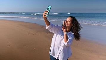 leende Lycklig lockigt håriga kvinna bloggare leende, inspelning video på henne smartphone, sändning luft kyss, talande förbi video länk, delning henne livsstil på social media, stående på hav sandig strand