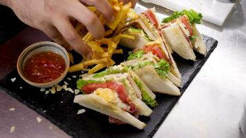 Cook hands placing fried french fries on a plate next to sandwiches in restaurant kitchen. Slow motion video