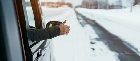 contento viajero conducción coche en Nevado la carretera y gesto dedo arriba, mujer turista disfrutando nieve bosque ver desde el coche ventana en invierno estación. invierno viajar, la carretera viaje, explorador y vacaciones conceptos foto