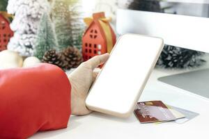 Gentle hands cradle a smartphone, empty screen by festive scene a beautiful Christmas tree adorned with credit cards, along with Christmas balls, pine cones, and red house gift box tied golden ribbon. photo