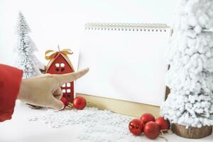 The finger is pointing at White calendar mock up to enter text by the festive scene of beautiful Christmas tree adorned, with Christmas balls, pine cones, red house gift box tied golden ribbon. photo