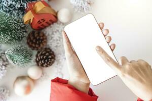 Top view Gentle hands cradle smartphone with a white screen mock and Fingers on the screen to shop online up by the festive scene of beautiful Christmas tree adorned, with Christmas balls, pine cones. photo