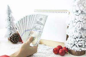 Gentle hands cradle money with a white screen mock-up and White calendar with a festive scene of a beautiful Christmas tree adorned, with Christmas balls, pine cones, and a red house gift box. photo