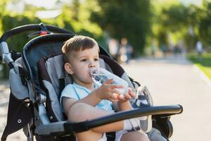 handsome boy sits in a stroller and drinks water from a bottle on a hot summer day photo