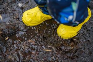 A little kid in yellow rubber boots is standing in the mud photo