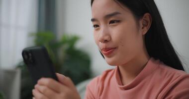 métrage de content Jeune asiatique femme se détendre sur confortable canapé à Accueil achats en ligne sur téléphone intelligent, souriant fille utilisation cellule téléphone bavardage, Feuilleter sans fil l'Internet sur gadget video