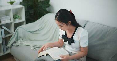 jovem ásia mulher sentado relaxante em sofá e lendo livro dentro vivo sala. video