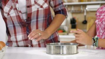 cerca arriba de joven hija tamiza harina mediante un tamiz a el cocina mientras su madre se sienta siguiente a su. lento movimiento imágenes video