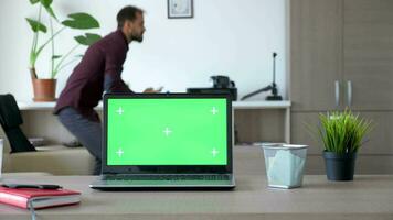 Laptop on the desk with a green screen in the house. A guy is walking in the background and sits on the couch looking at the smartphone video