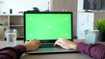 Male hands typing on the laptop with a mock up green screen chroma in the living room. He is in his apartment at the desk. POV, dolly slider 4K footage video