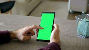 Male hands scrolling on smartphone with green screen chroma mock up at his desk in the living room of his home. Dolly revealing slider 4K footage video