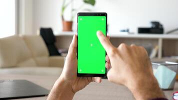 Mens hand- scrollen naar beneden en Rechtsaf Aan een modern smartphone met groen scherm chroma bespotten omhoog in zijn comfortabel leven kamer Aan de achtergrond video