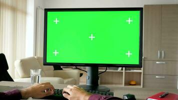 först person se man händer skriver på dator tangentbord med stor grön skärm krom mockup. de pc är på de skrivbord i levande rum och de TV är på i de bakgrund. dolly reglaget 4k antal fot med parallax effekt video