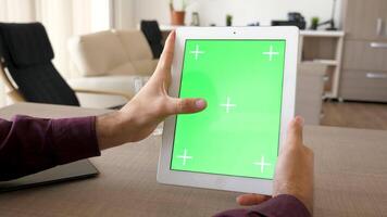 Tablet PC in vertical position with green screen chroma mockup beeing hold by male hands on the desk in house video