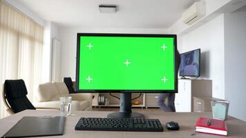 Personal PC computer with big green screen chroma mock up on the table in the living room. A guy is entering the room in background while the TV is on and sits on the sofa looking at his phone video