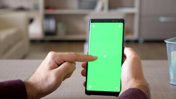 vicino su di uomo navigazione il Internet su smartphone con verde schermo croma finto su a il suo scrivania nel il vivente camera video