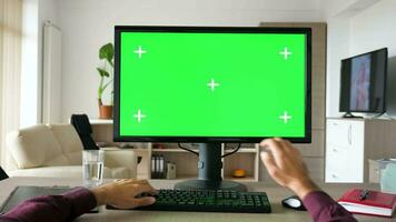 First person view man hands typing on computer keyboard with big green screen chroma mockup. The PC is on the desk in living room and the TV is on in the background video