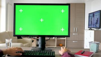 First person view man hands typing on computer keyboard with big green screen chroma mockup. The PC is on the desk in living room and the TV is on in the background. Sliding from right to left 4K footage video