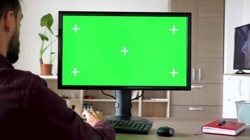 Side view of man sitting in front of computer with big green screen mockup chroma typing on the keyboard. He is in the living room of his house video