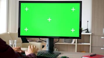 Side view of man sitting in front of computer with big green screen mockup chroma typing on the keyboard. He is in the living room of his house. Dolly slider 4K footage with parallax effect video