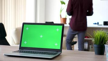 Laptop on a desk in the living room with a isolated green screen and marks for tracking. Man walks in the room and sits on the couch in the background video