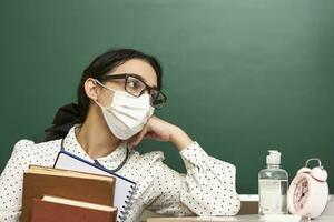 a young teacher wearing a face mask sits at a desk with books photo
