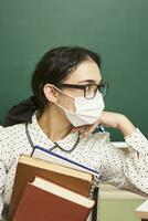 a young teacher wearing a face mask sits at a desk with books photo
