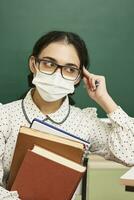 a young teacher wearing a face mask sits at a desk with books photo